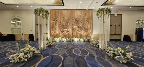 Wide view of a sculpted champagne fan-pleat stage backdrop edged with white and blue florals, flanked by crystal-strand pillars topped with floral arrangements.