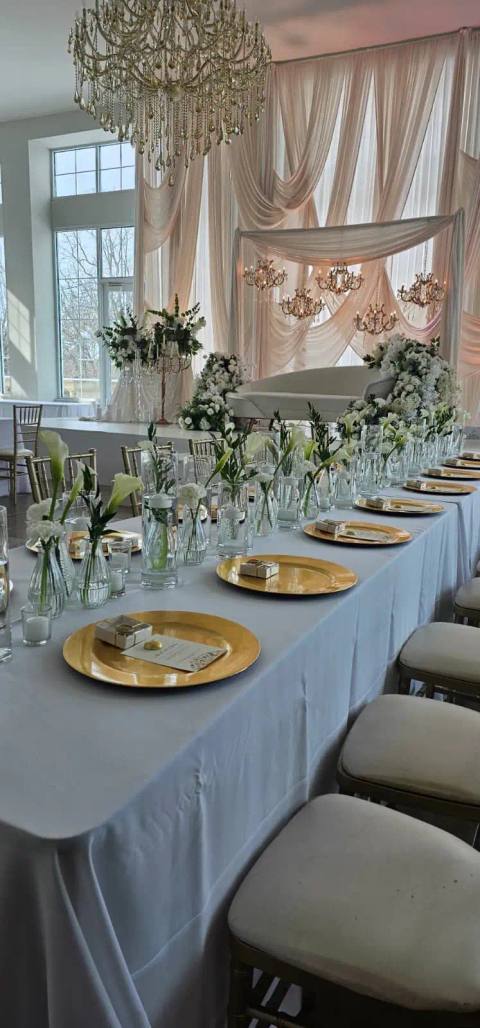 Reception hall with a long head table set with gold chargers and calla lilies, blush drapery and chandeliers across the stage, and a crystal chandelier overhead.