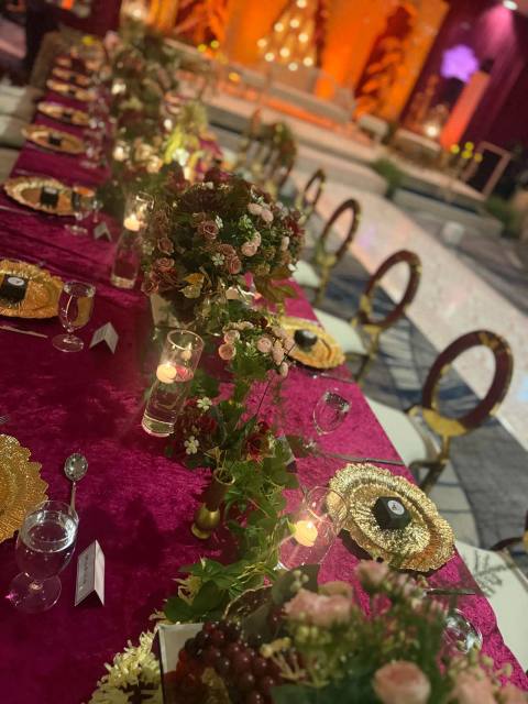 Long table in magenta crushed velvet with gold chargers, candles in glass, garden roses and trailing greenery, gold ring-back chairs and a warmly lit stage behind.