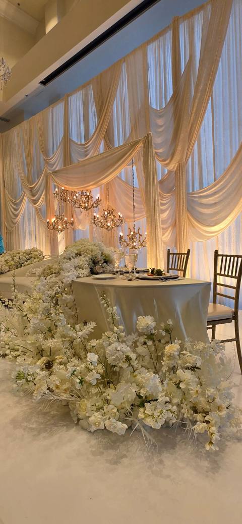 Sweetheart table in ivory linen with a low bank of white flowers spilling across the floor, before warmly lit ivory swag drapery hung with crystal chandeliers.