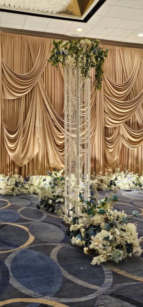 Champagne swag drapery behind a crystal-strand pillar crowned with white and blue florals, with a low line of flowers along the stage edge.