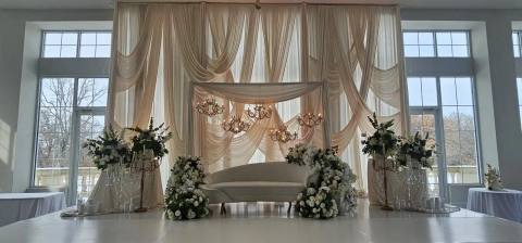 Reception stage in a bright hall with tall windows: ivory swag drapery, crystal chandeliers, a white curved sofa, white rose arrangements and gold candelabras.