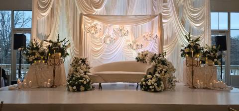 Ivory drape stage lit warm with candles, a white curved sofa between banks of white roses, and gold candelabras dressed with greenery.
