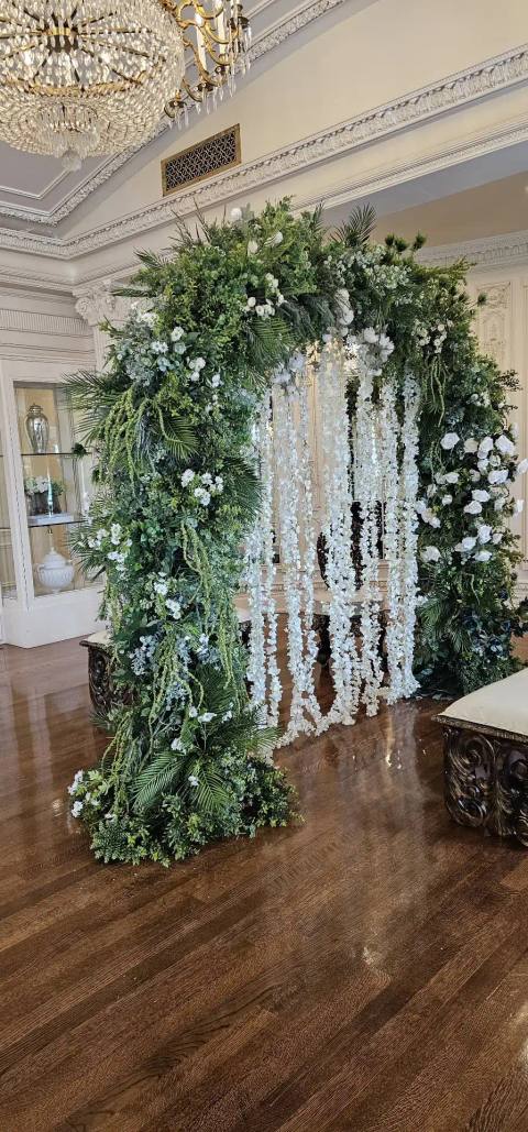 Ceremony arch covered in lush greenery and white blooms, hung with strands of white wisteria, in a white paneled room with a chandelier.