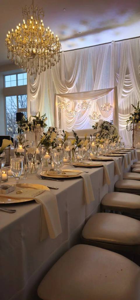 Long reception table with gold chargers, folded ivory napkins, candles and low white florals, under a gold crystal chandelier with ivory drapery beyond.