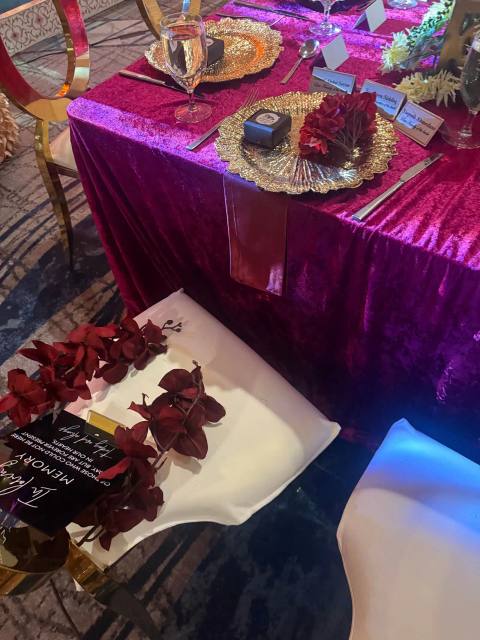 A reserved chair draped with burgundy orchids and a memorial sign, beside a magenta velvet table set with gold chargers, crystal glassware and place cards.