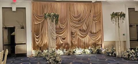Champagne swag drape backdrop with two crystal-strand pillars crowned in white florals and a line of white and blue flowers along its base.
