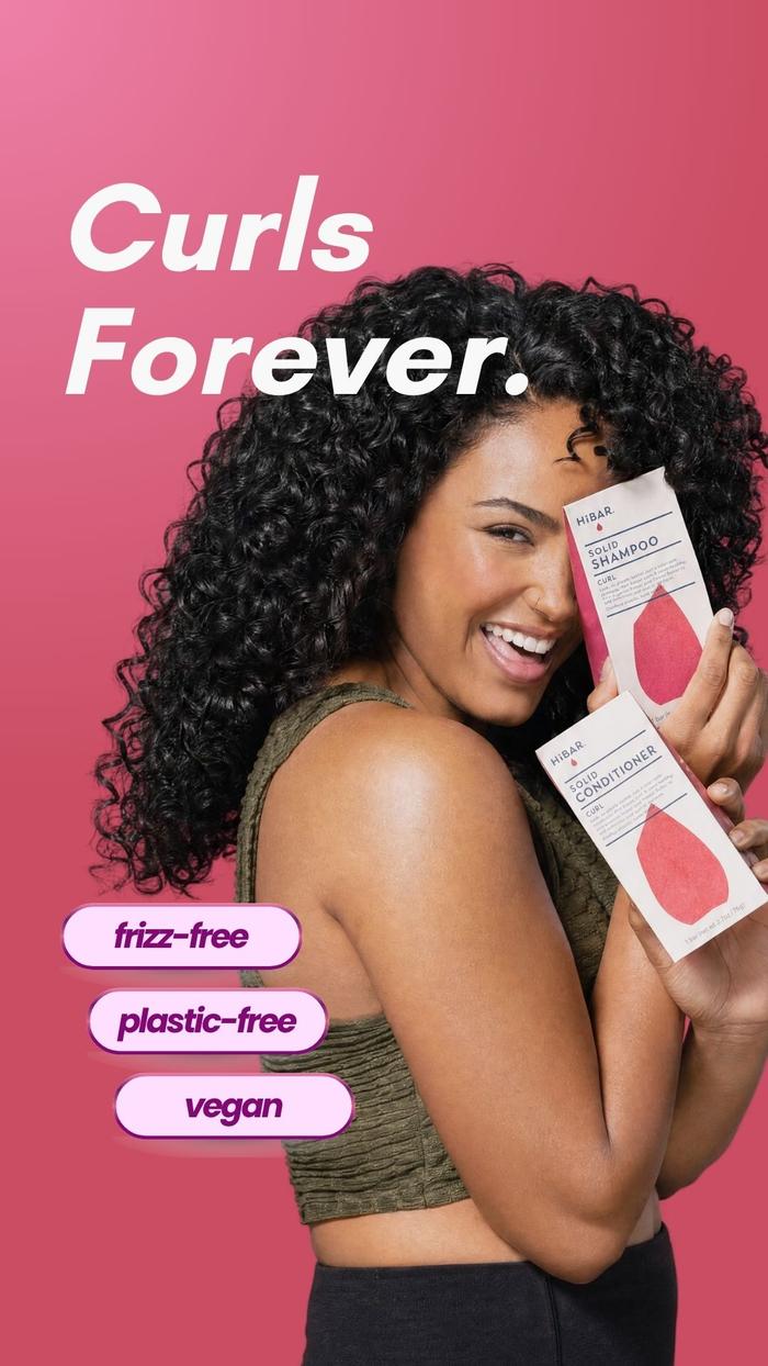 Curly haired woman holding up HiBAR shampoo and conditioner for curly hair against pink background.