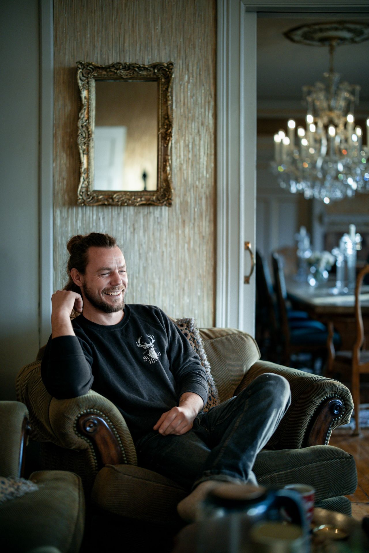 Peder Bolstad sitter i en lenestol og ler, med spisestue og lysekrone i bakgrunnen.