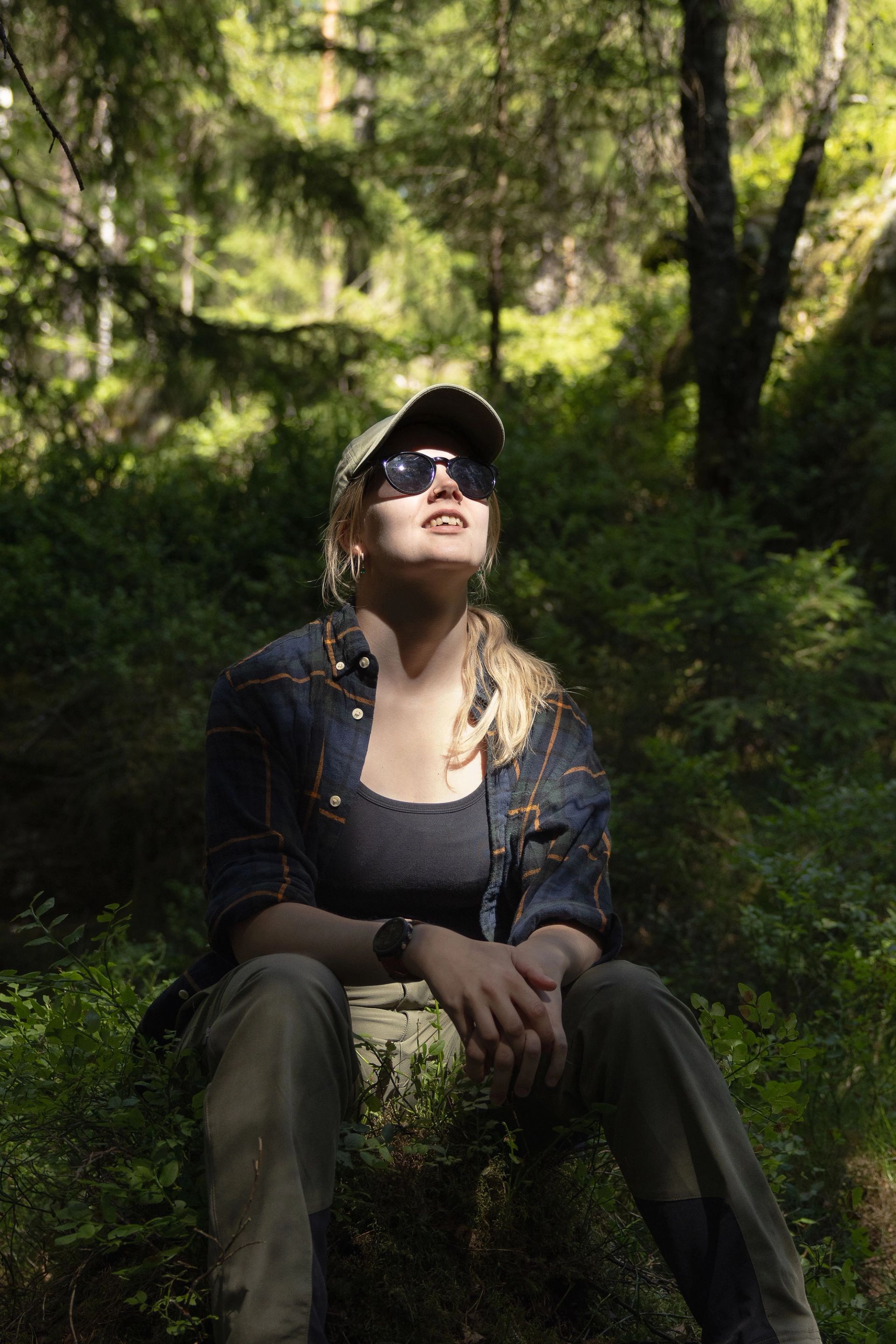 Elin Johanne sitter på skogbunnen med solbriller og ser opp i skogen.