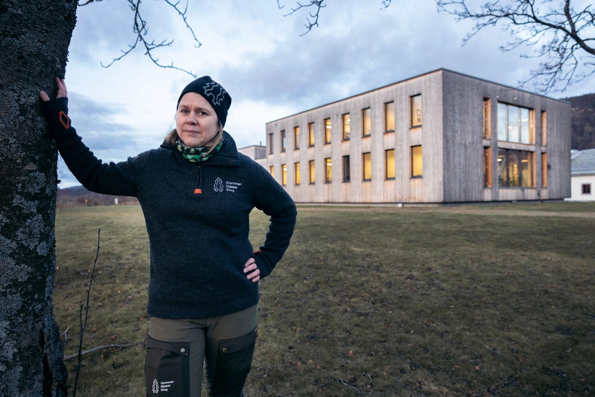 Ane Christensen Tange står ved et tre foran en moderne trebygning.