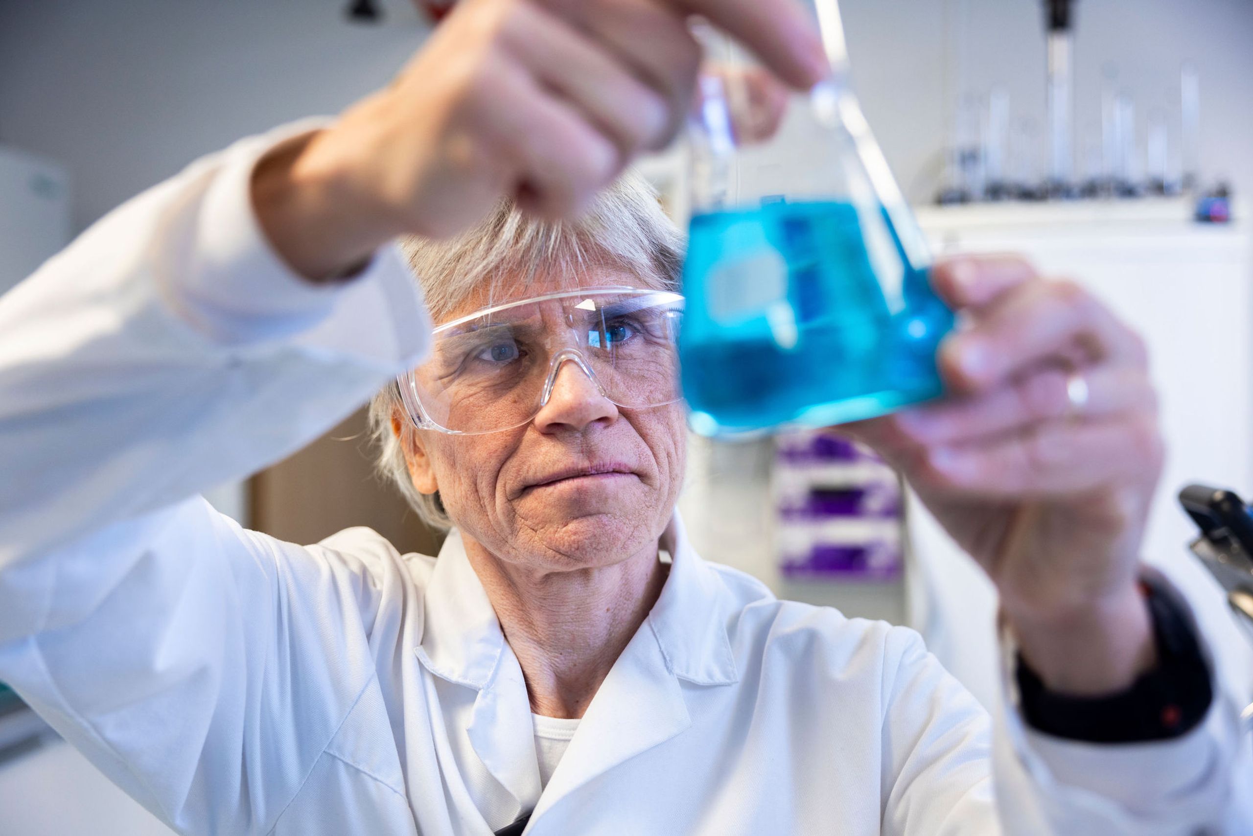 Kristin Misund i hvit laboratoriefrakk og vernebriller undersøker en kolbe med blå væske.