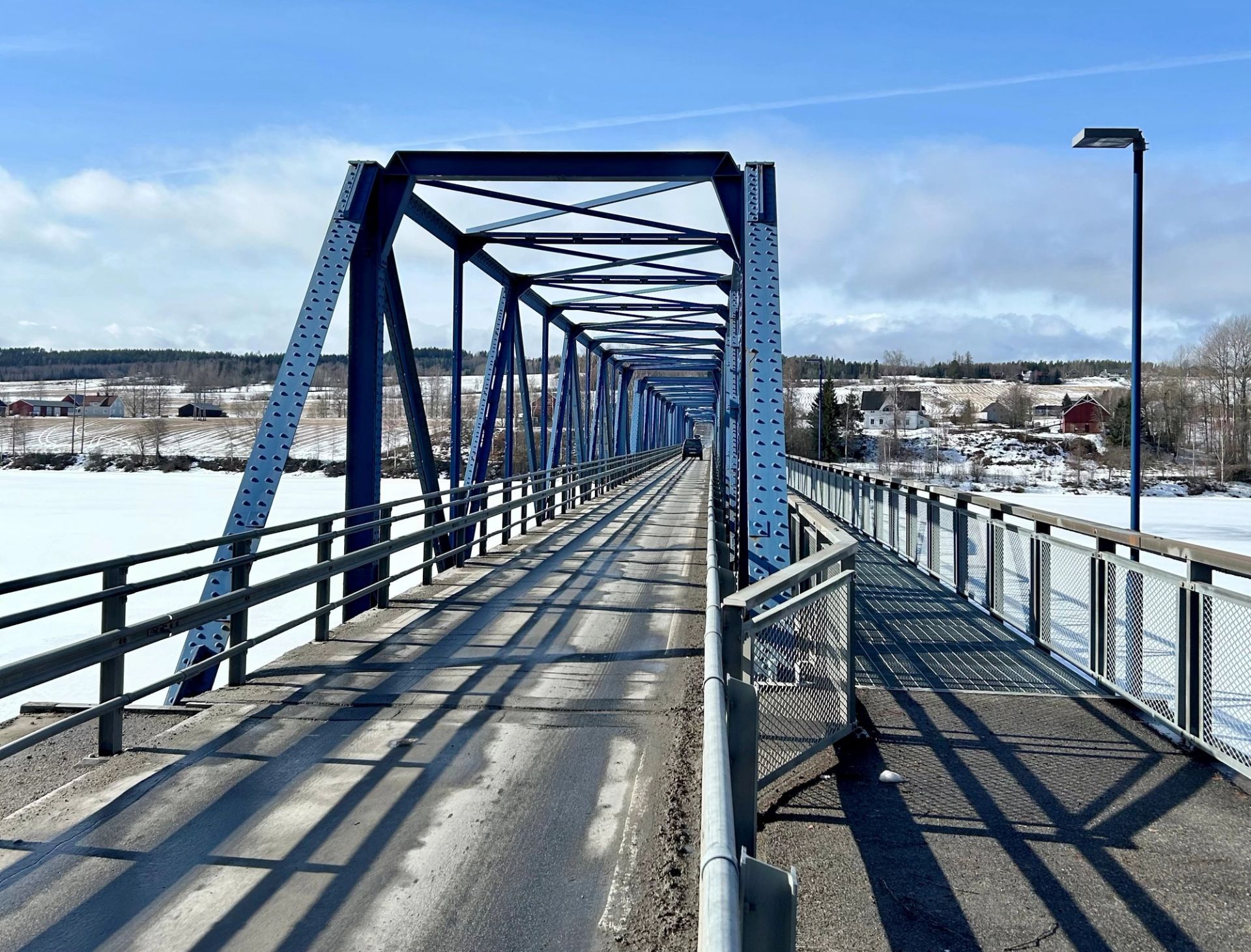 Blå stålbru over en islagt elv, med snødekte jorder og bebyggelse i bakgrunnen.