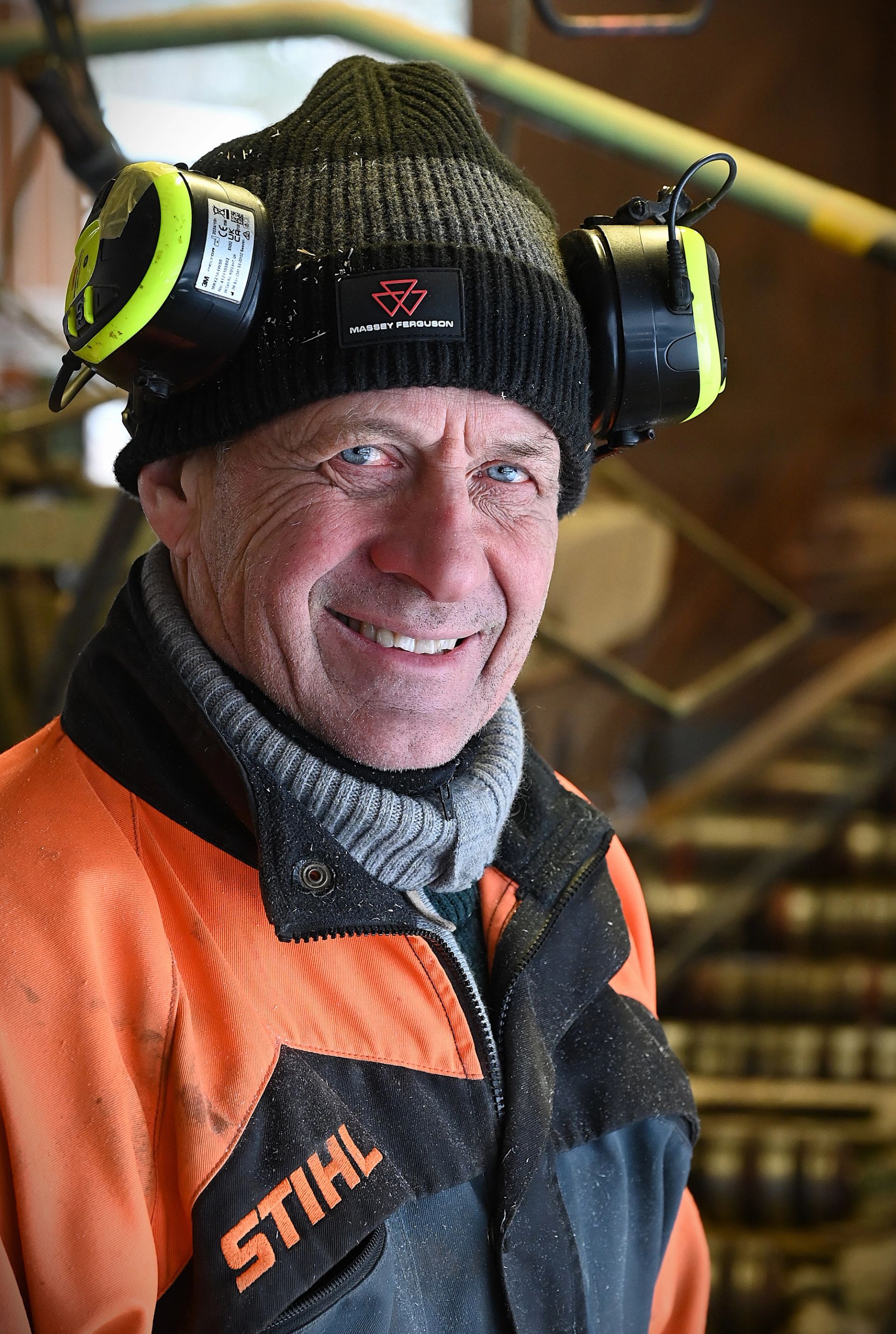 Runar Bekkeseth smiler mot kamera iført svart lue med hørselvern og oransje arbeidsjakke inne på sagbruket.