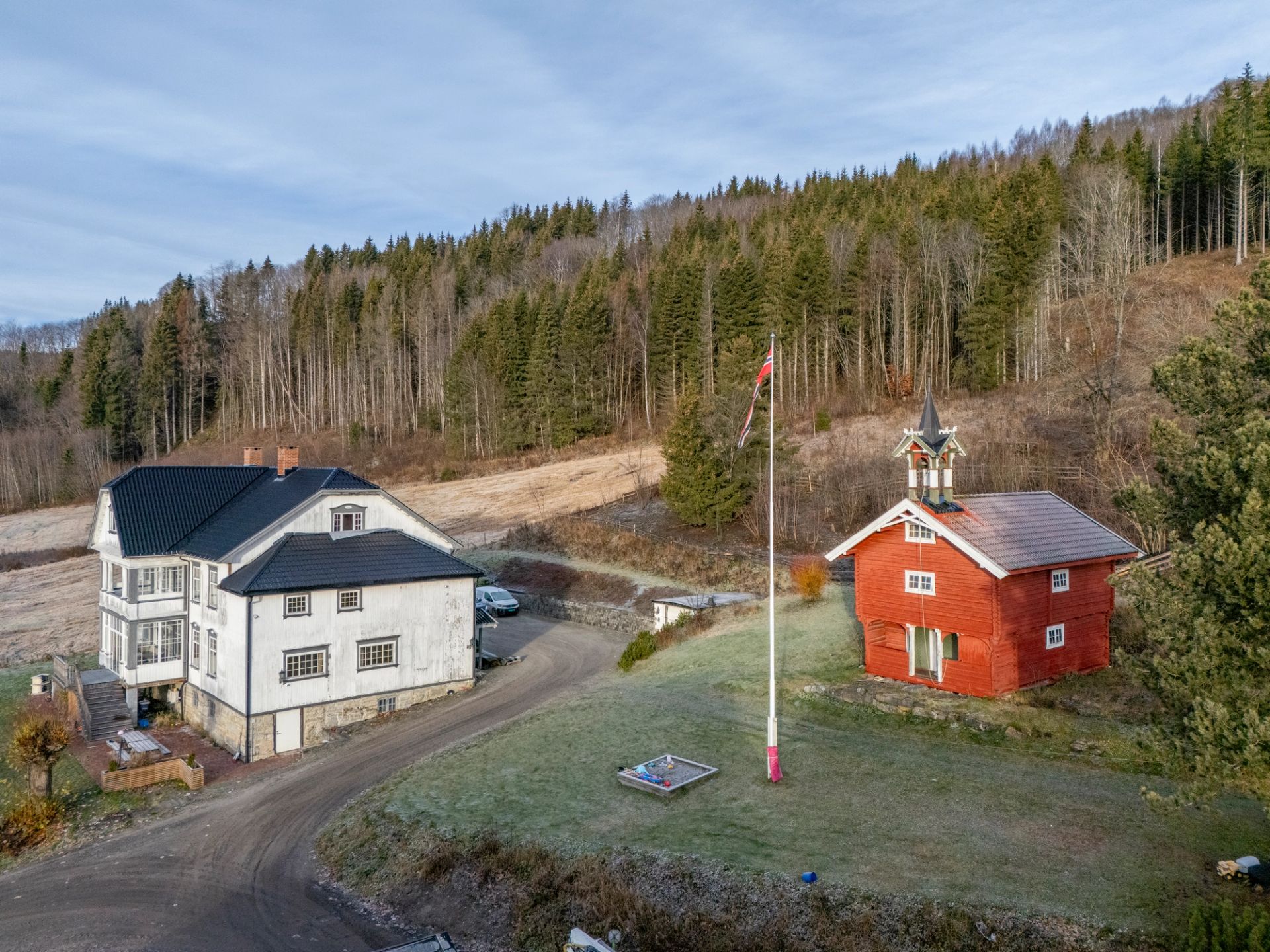 Gårdstun med hvitt våningshus, rødt stabbur og norsk flagg, omgitt av skogkledde åser.
