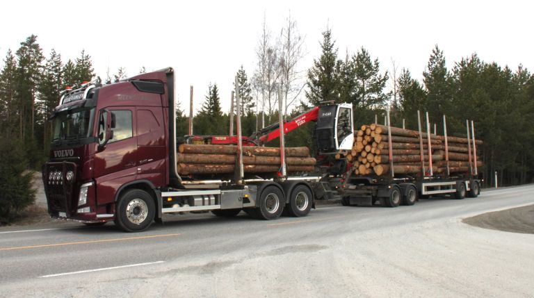Rødt tømmervogntog med kran og tømmer på lasteplanet og tilhengeren. Vogntoget er ikke fullastet og står på en vei omgitt av skog.