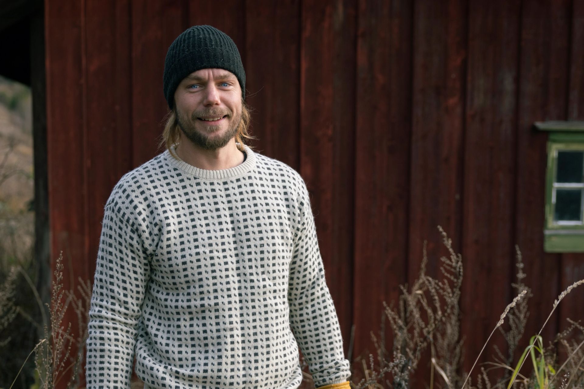 Peder Bolstad står foran en rød trebygning iført lue og strikkegenser.