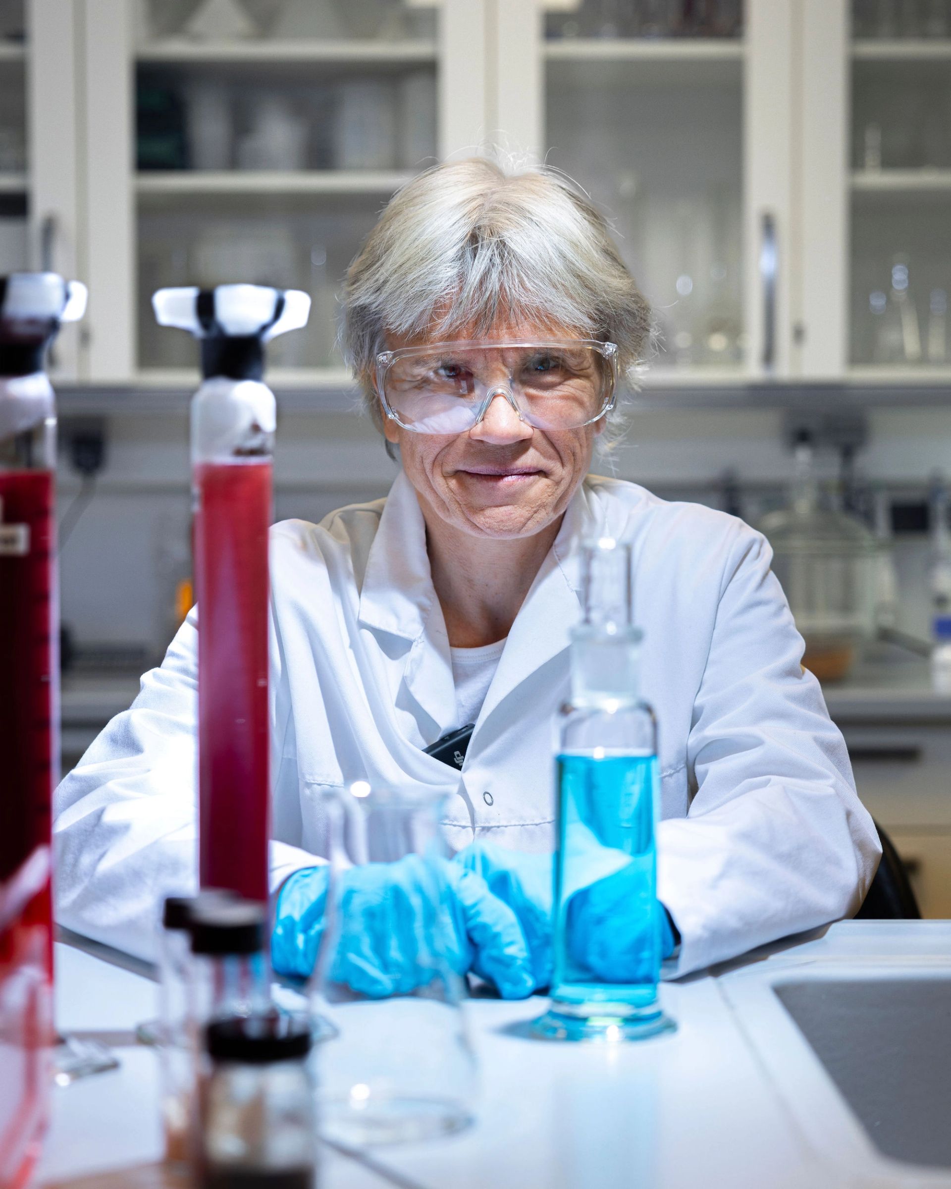 Kristin Misund med laboratoriefrakk, vernebriller og blå hansker sitter ved en laboratoriebenk med røde og blå væsker.