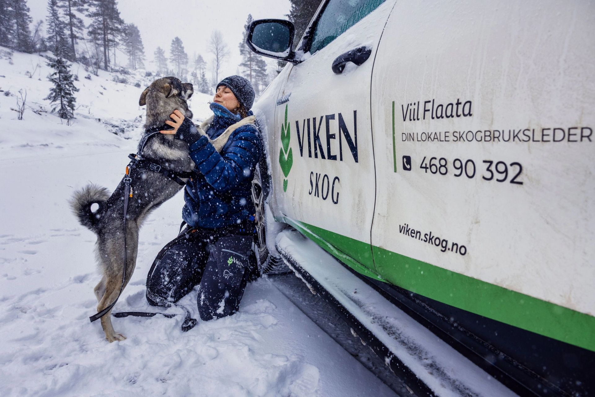 Viil Flaata sitter på kne i snøen og hilser på hunden sin ved en bil fra Viken Skog.
