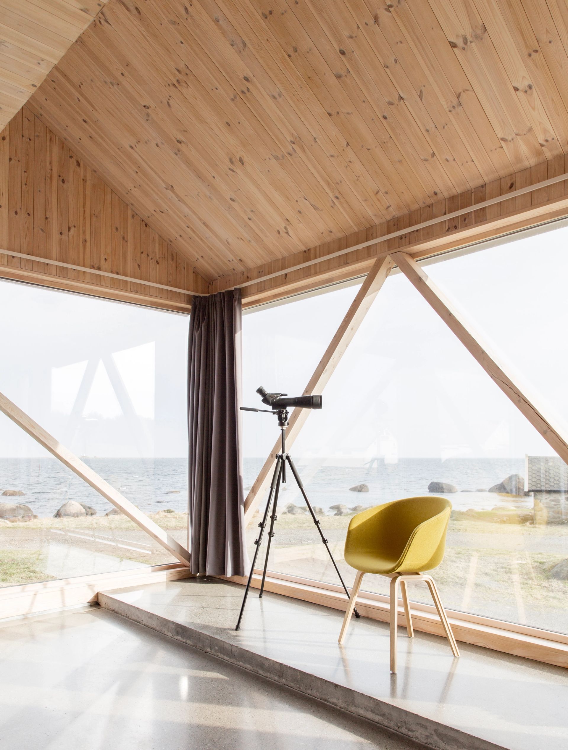 Interiør med trepanel, store panoramavinduer mot havet, stol og teleskop.