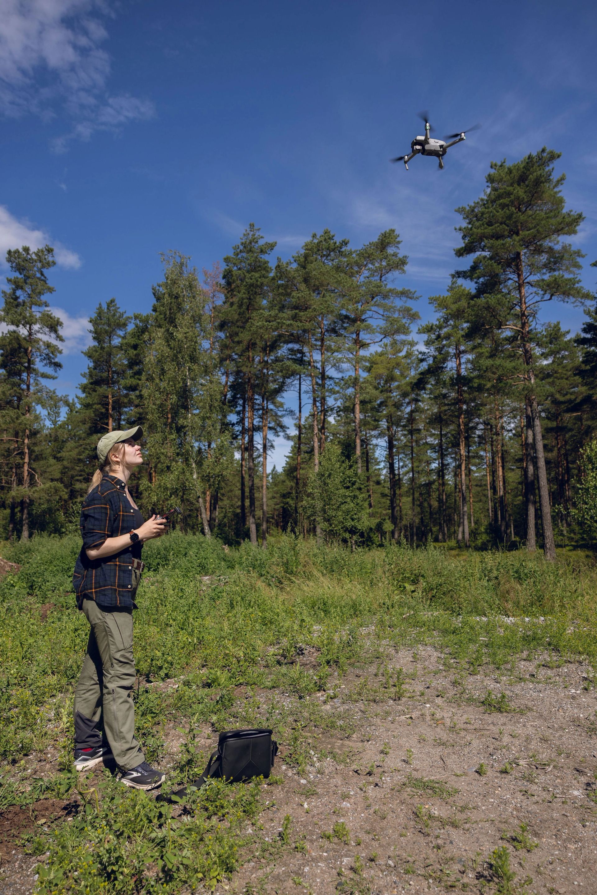 Kvinne styrer en drone som flyr over en åpen plass i skogen.