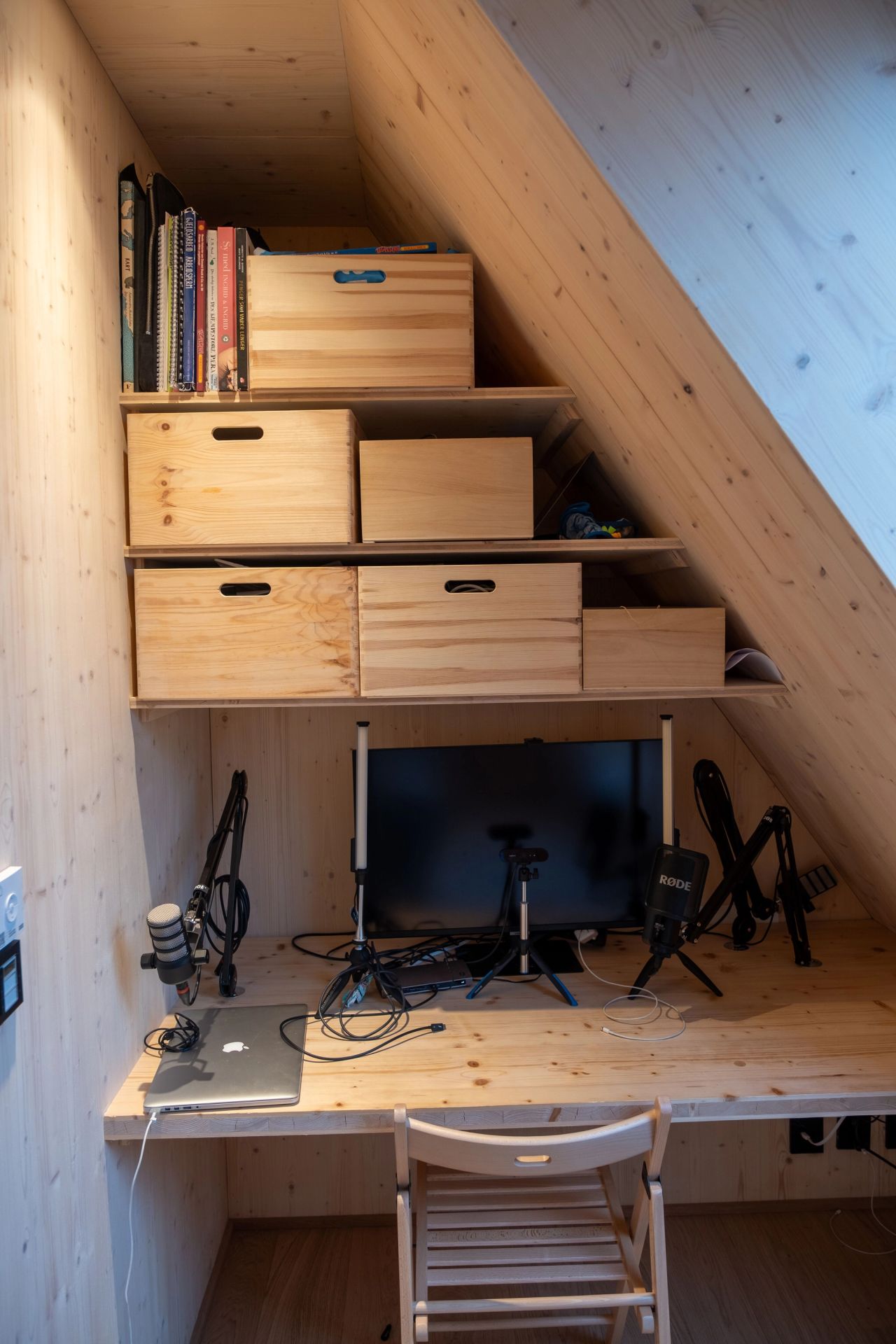 Lite hjemmekontor under skråtak, med skrivebord, skjerm, mikrofoner og oppbevaring i tre.