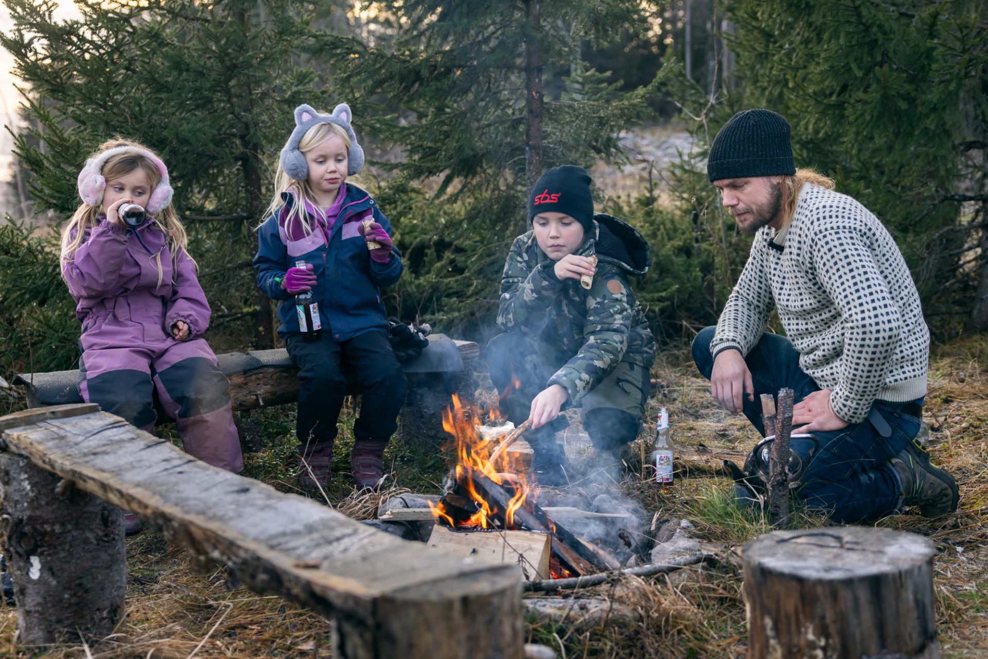Peder Bolstad sitter ved et bål i skogen sammen med tre barn som spiser og drikker.