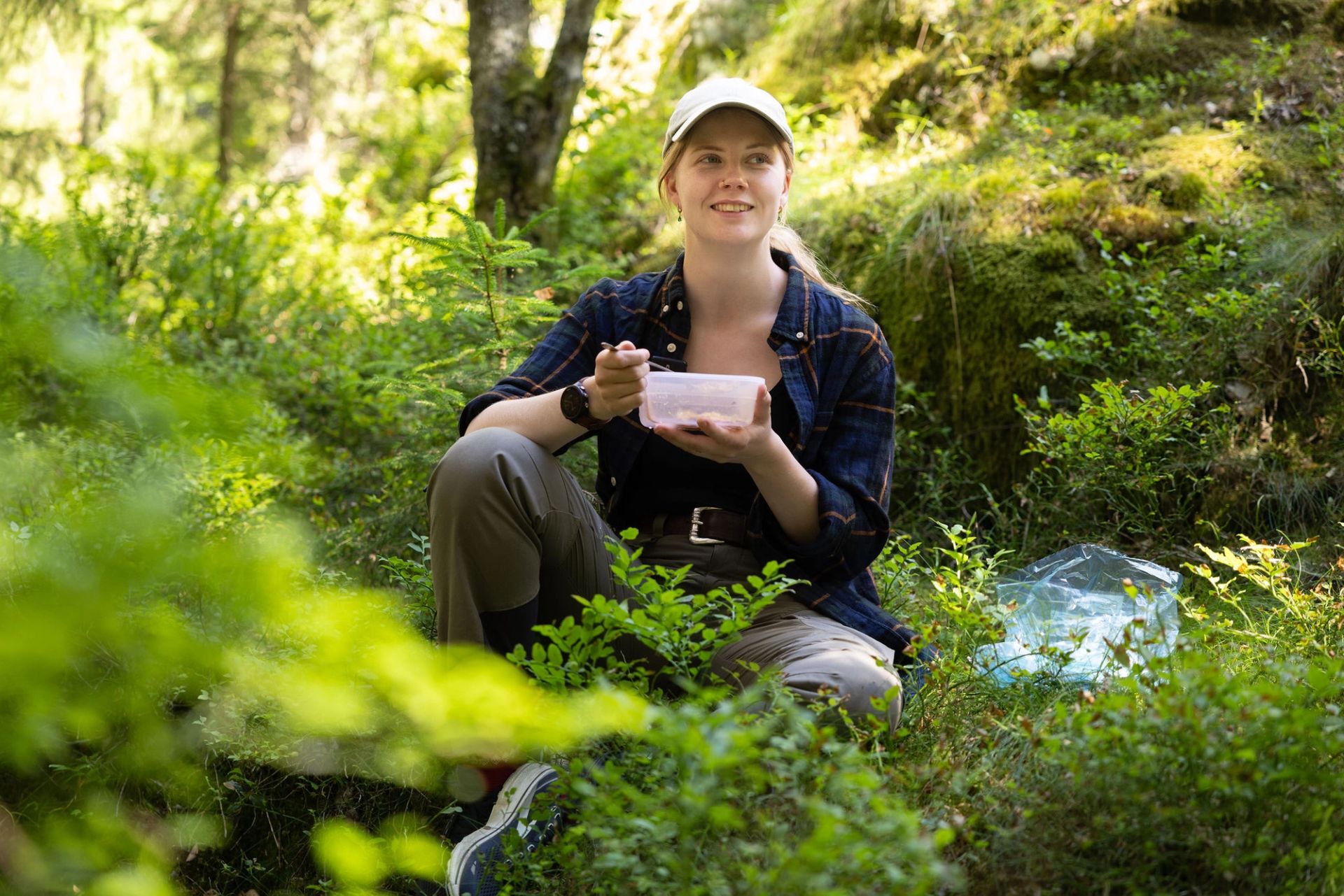 Elin Johanne sitter på skogbunnen og spiser mat fra en plastboks.