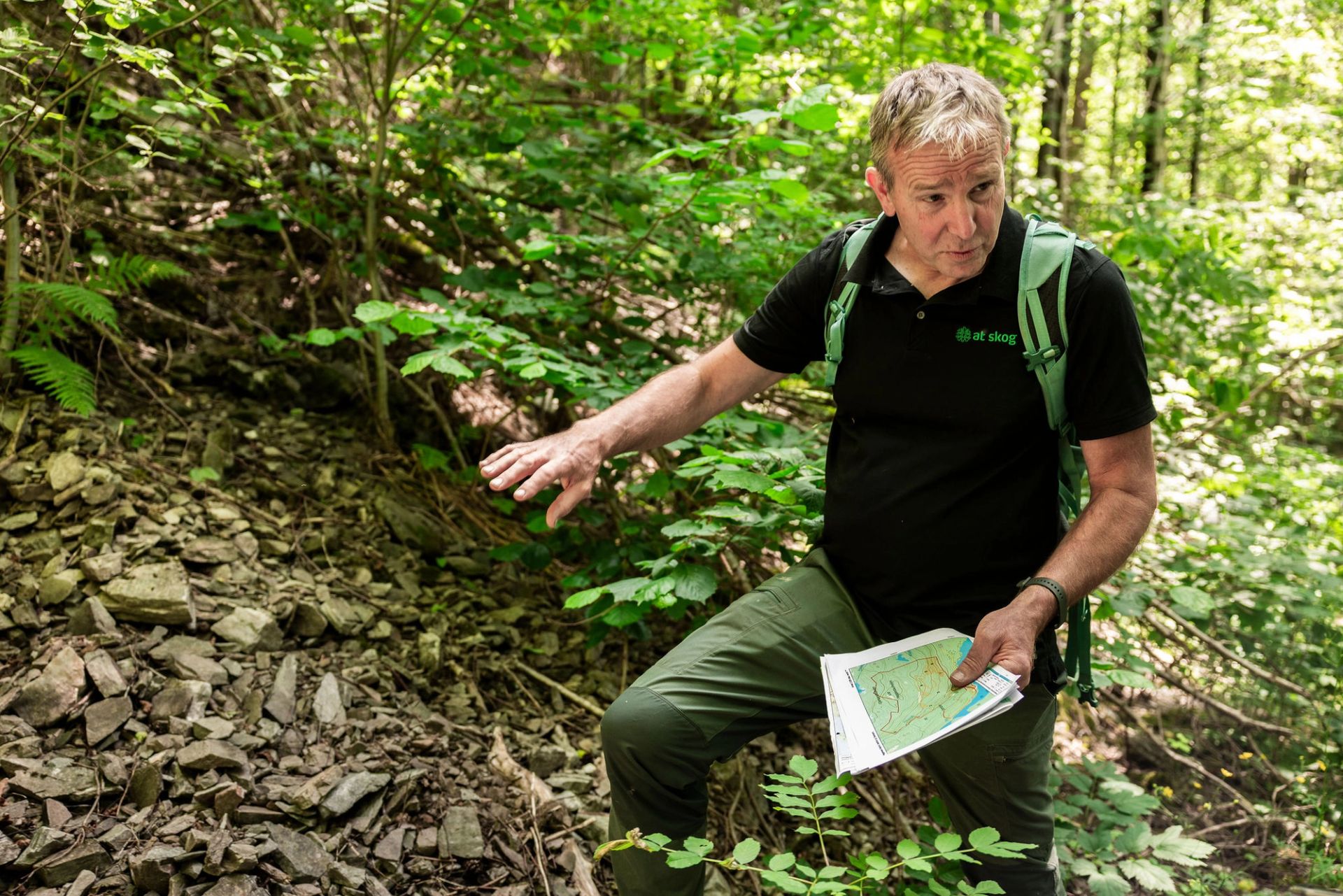 Mann i skogen peker mot stein og vegetasjon i en bratt skråning.