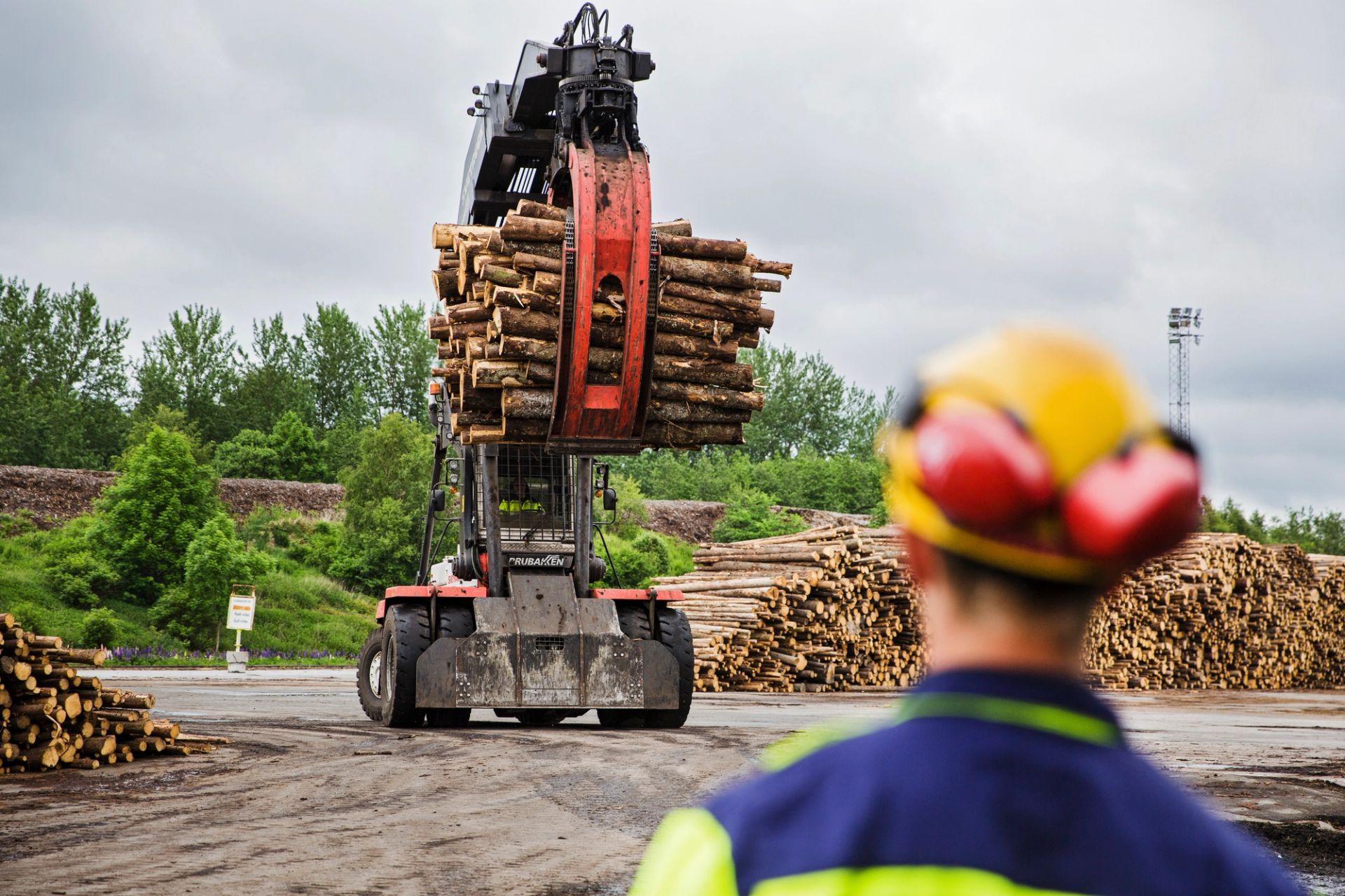 Truck løfter en bunt med tømmer på et industriområde, mens en arbeider med hjelm står i forgrunnen.