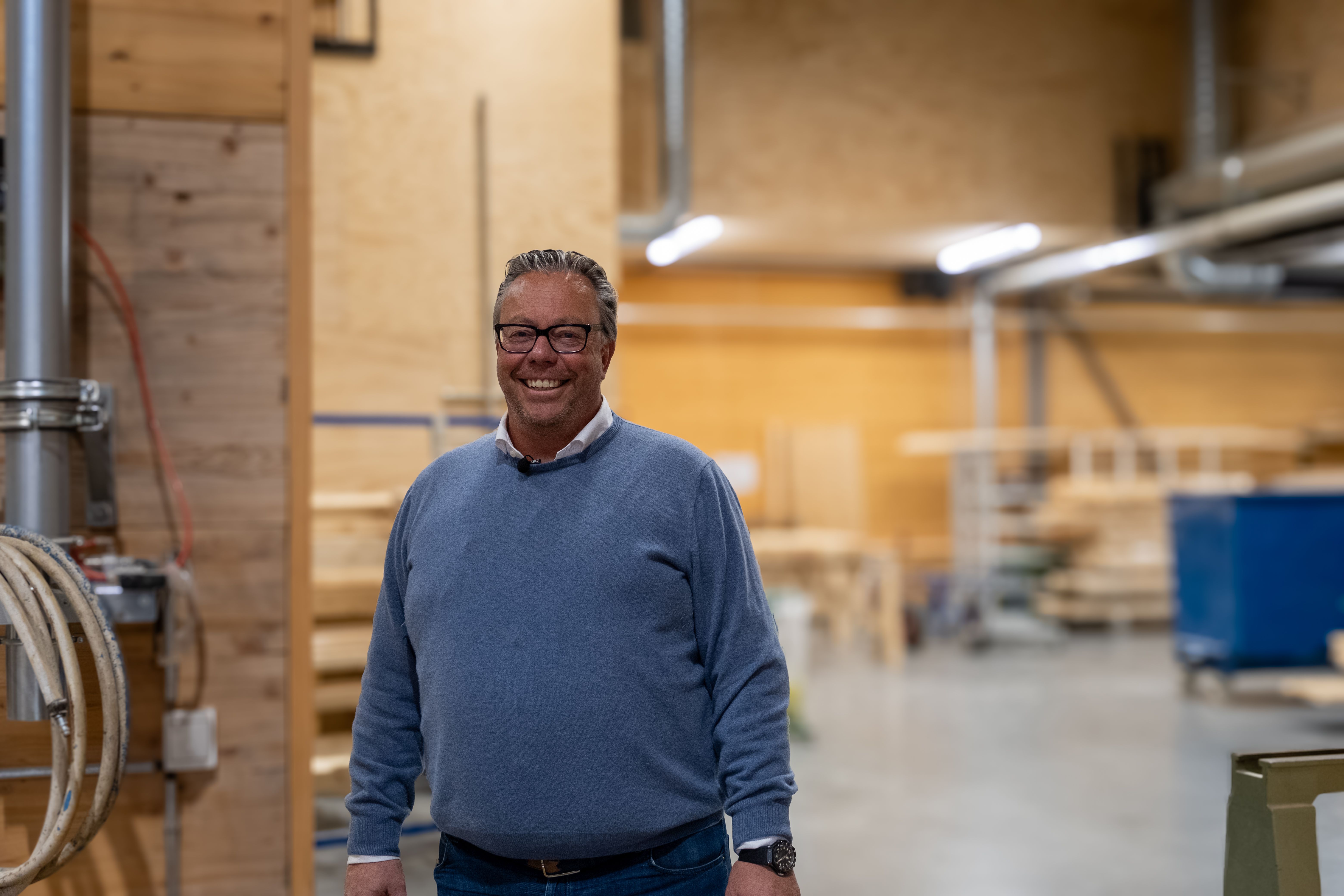 Gunnar-Adolf Aanesland smiler i fabrikken, omgitt av trekonstruksjoner og produksjonsutstyr.