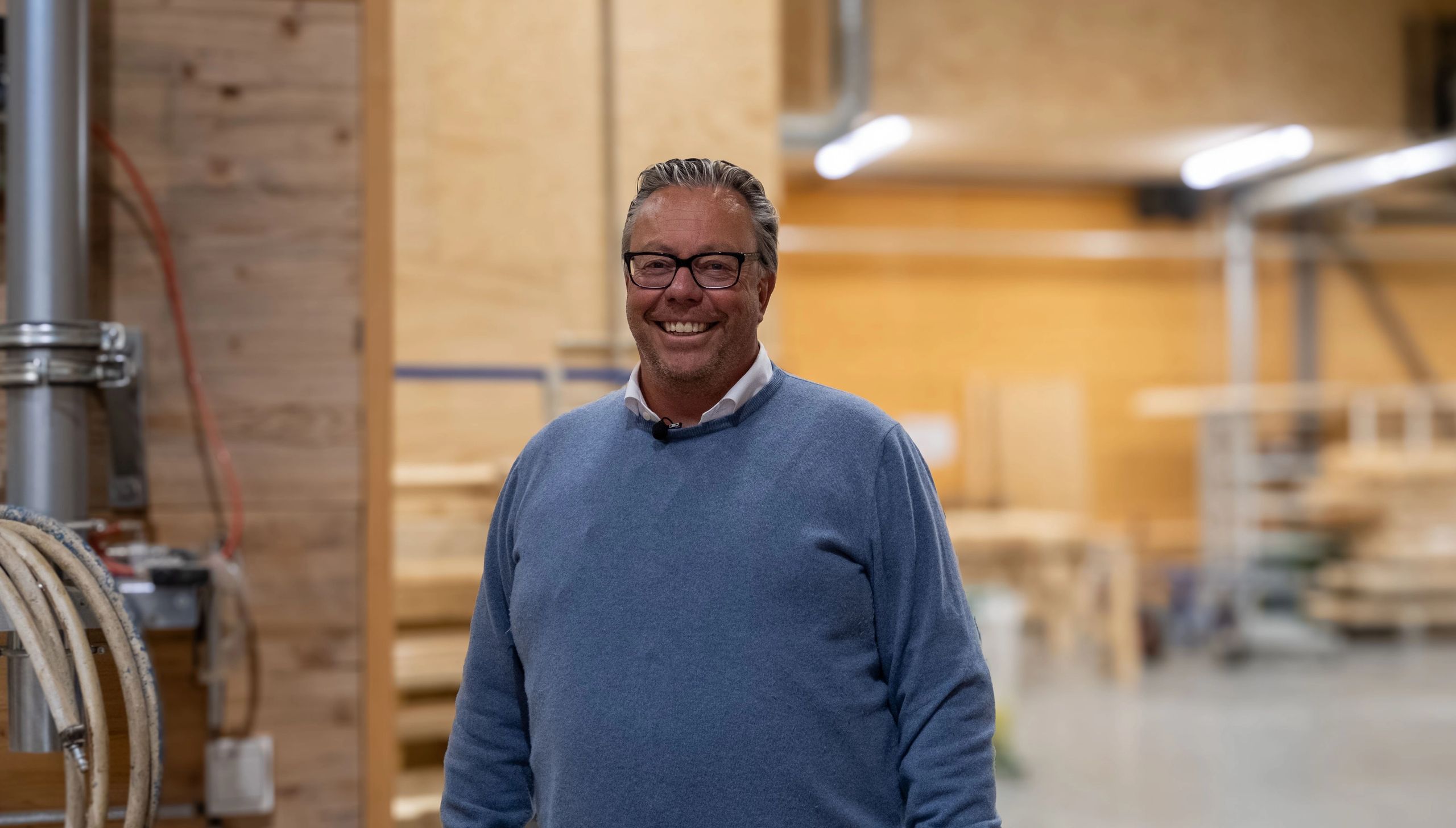 Gunnar-Adolf Aanesland smiler i fabrikken, omgitt av trekonstruksjoner og produksjonsutstyr.