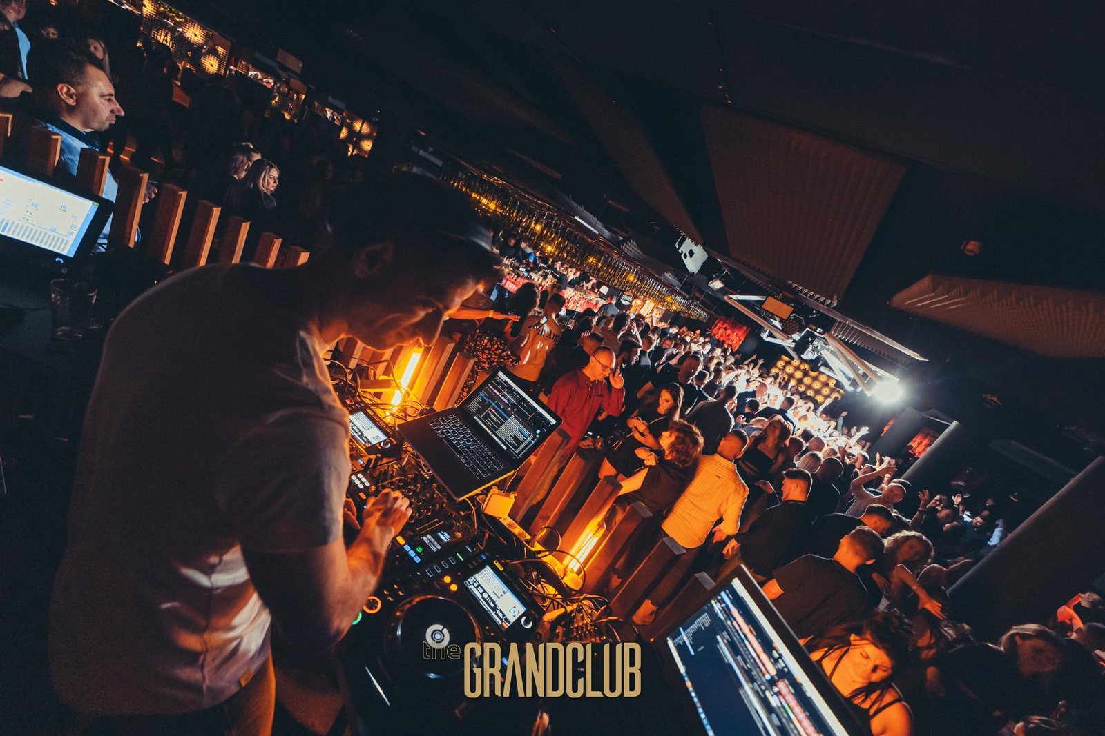 A view from behind the DJ booth showing the artist performing at a modern mixing console, overlooking a long, vibrant bar area packed with guests under warm orange lighting.