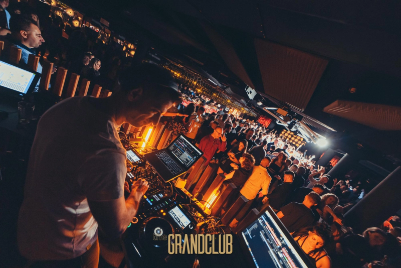 A view from behind the DJ booth showing the artist performing at a modern mixing console, overlooking a long, vibrant bar area packed with guests under warm orange lighting.