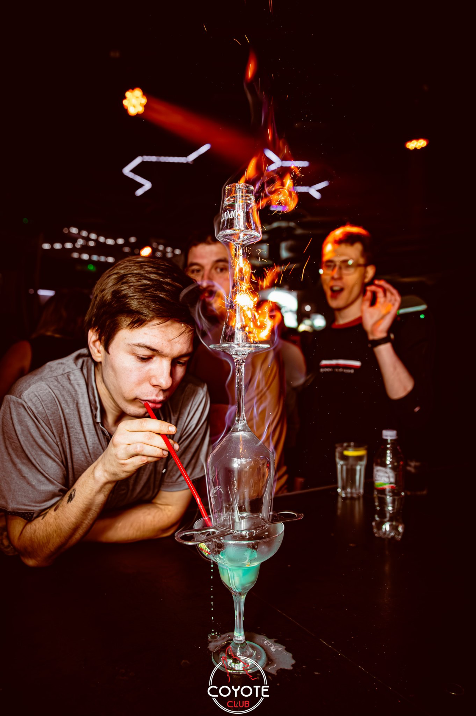 A high-angle shot of a guest drinking a cocktail through a straw from a flaming tower of stacked glasses at the bar, with other guests cheering in the background.