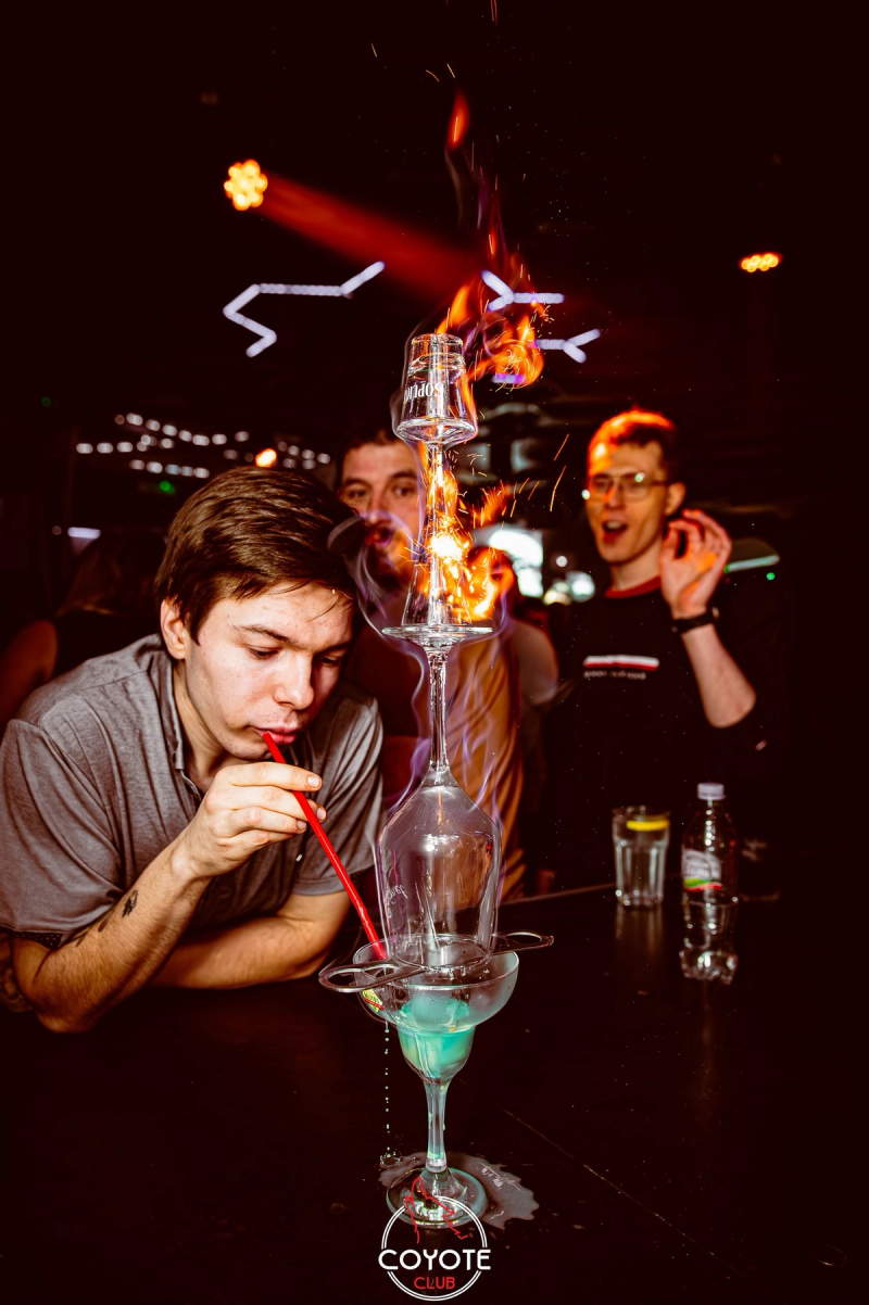 A high-angle shot of a guest drinking a cocktail through a straw from a flaming tower of stacked glasses at the bar, with other guests cheering in the background.