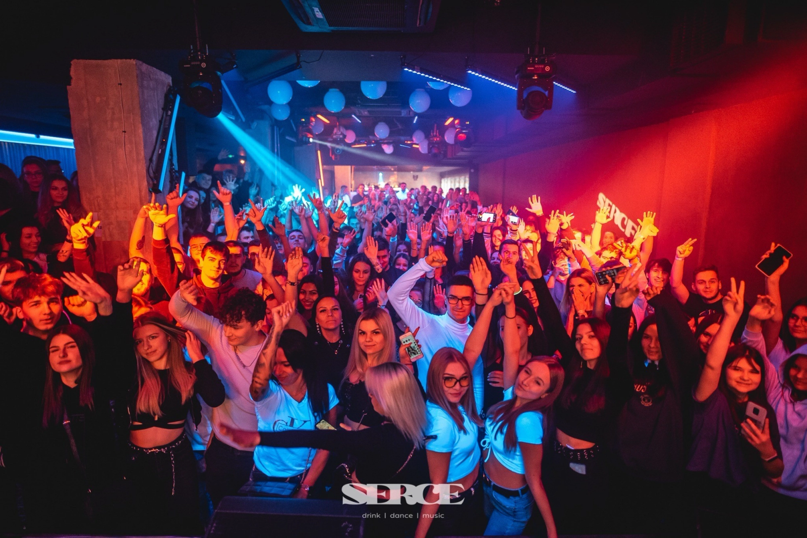 A large, energetic crowd with their hands raised at SERCE Lublin nightclub, illuminated by vibrant red and blue spotlights.