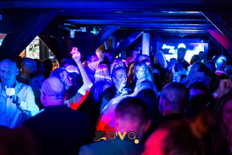 A crowded dance floor at AVA 3.0 bathed in deep blue and red lighting, with guests dancing under a low-ceilinged, industrial-style interior.