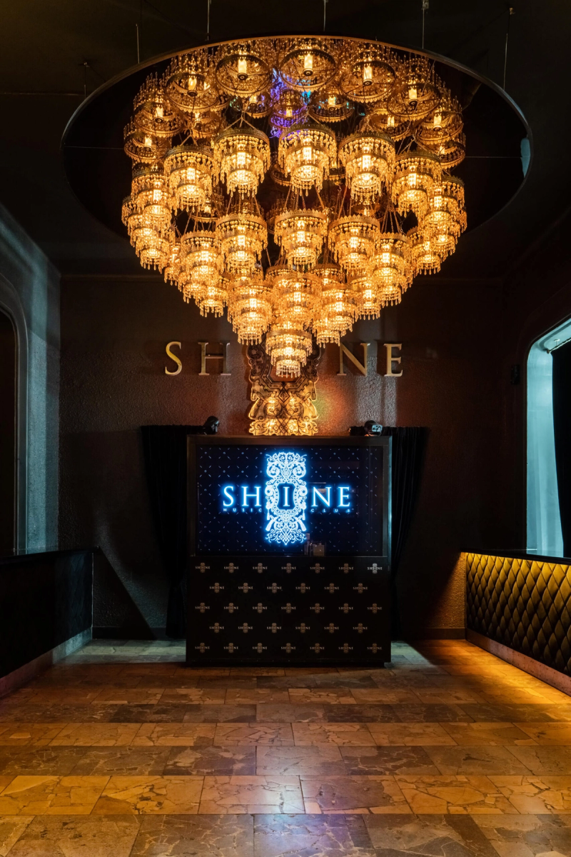 Grand entrance of Shine Club Kraków featuring a massive, multi-tiered crystal chandelier illuminating a lobby with marble floors and a digital screen displaying the Shine Music Club logo.