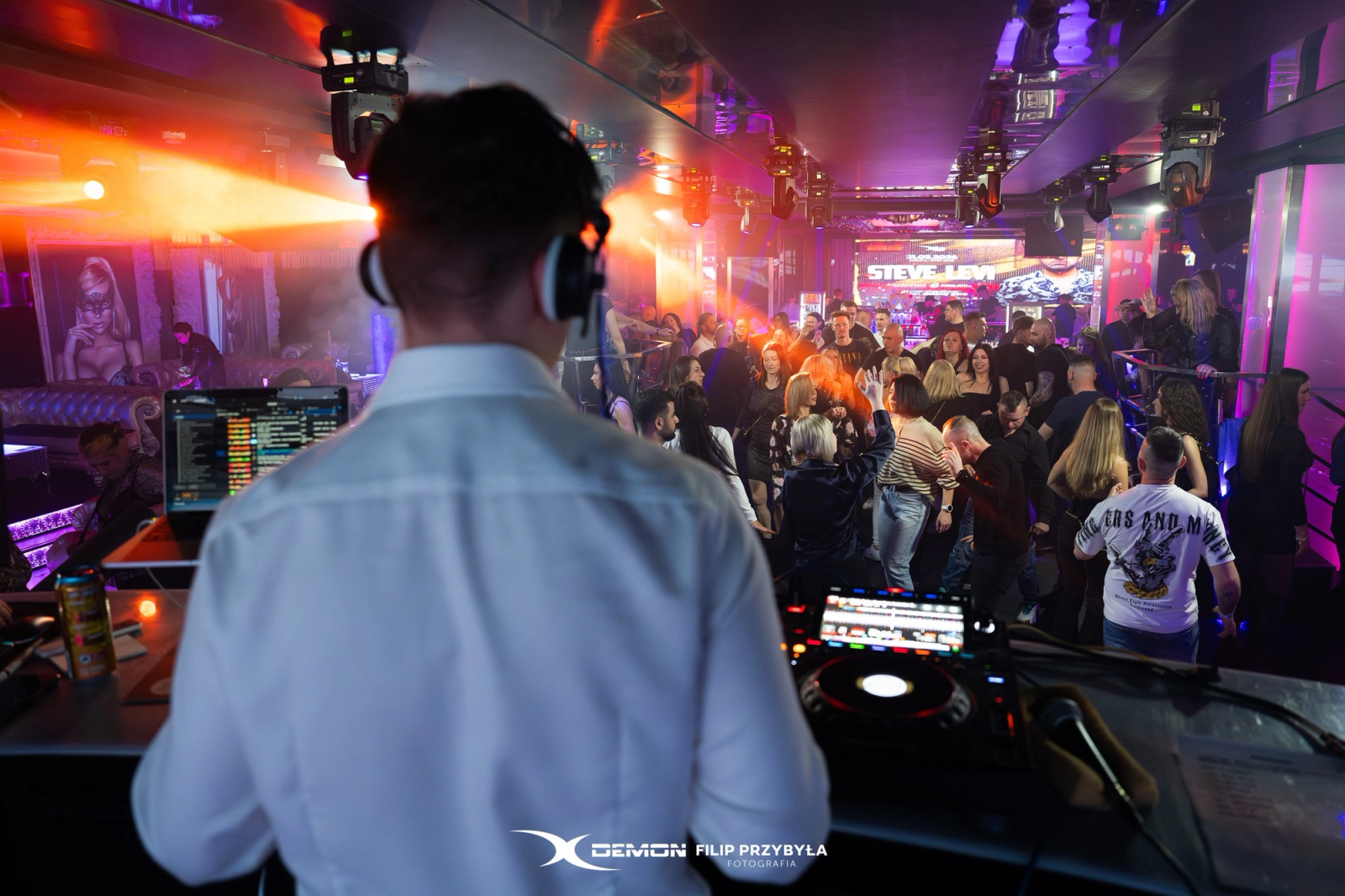An over-the-shoulder view of a DJ in a white shirt and headphones performing for a crowded dance floor at X-Demon Zielona Góra, illuminated by warm orange spotlights.
