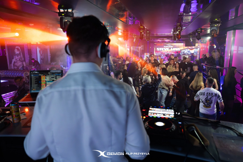 An over-the-shoulder view of a DJ in a white shirt and headphones performing for a crowded dance floor at X-Demon Zielona Góra, illuminated by warm orange spotlights.