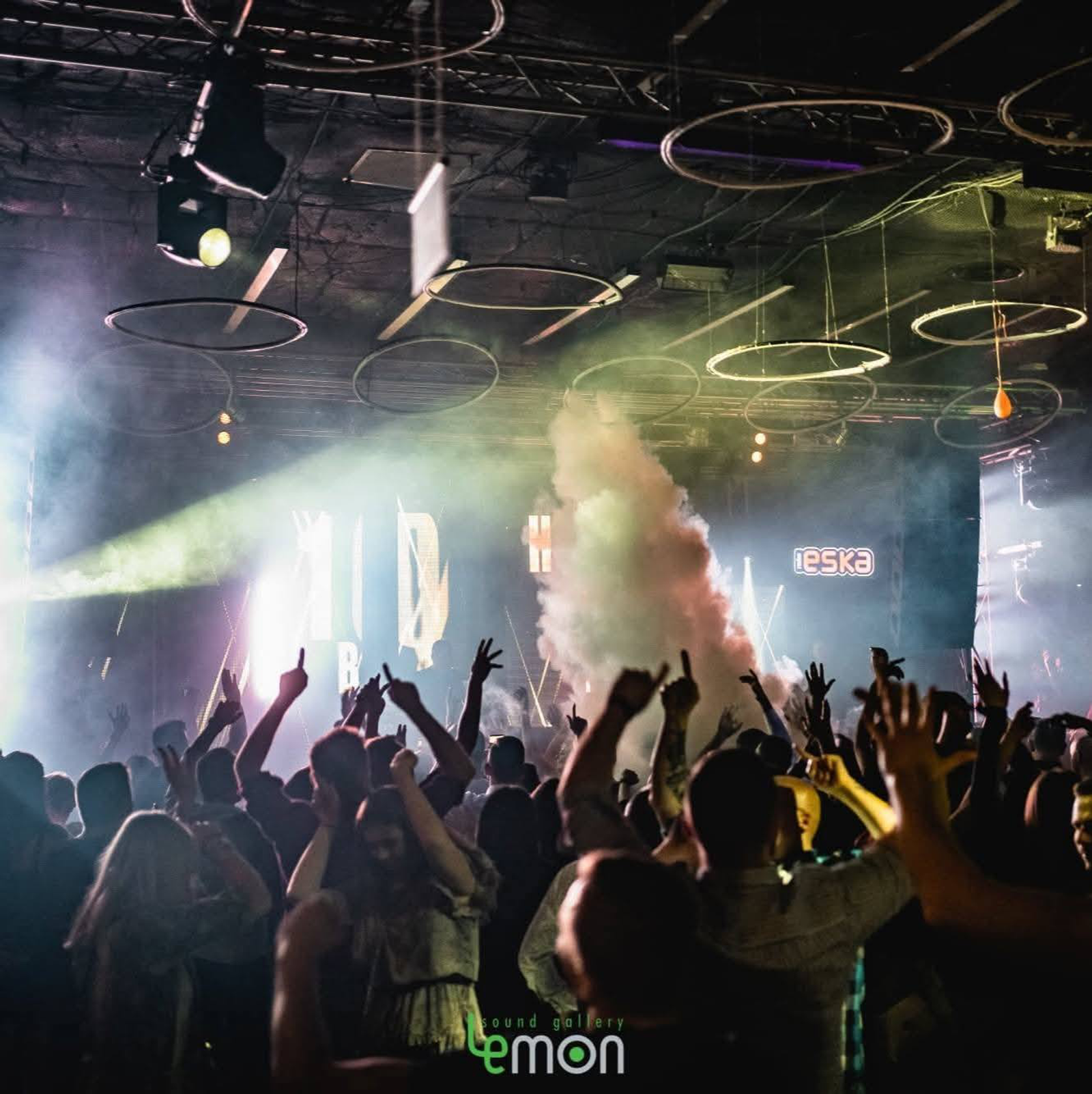 An energetic crowd at LEMON Sound Gallery with hands raised, illuminated by stage lights and a dramatic smoke blast. Large glowing circular rings hang from the industrial ceiling, and a screen displays the Radio Eska logo.