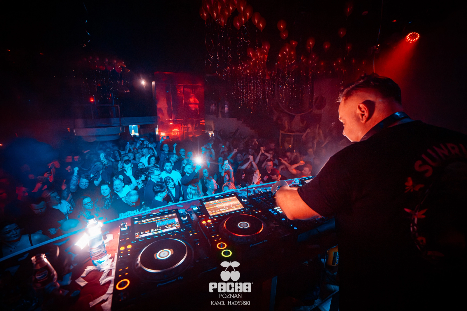 A view from behind the DJ booth at Pacha Poznań, showing the artist's hands on the decks overlooking a massive crowd under red atmospheric lighting and ceiling balloons.