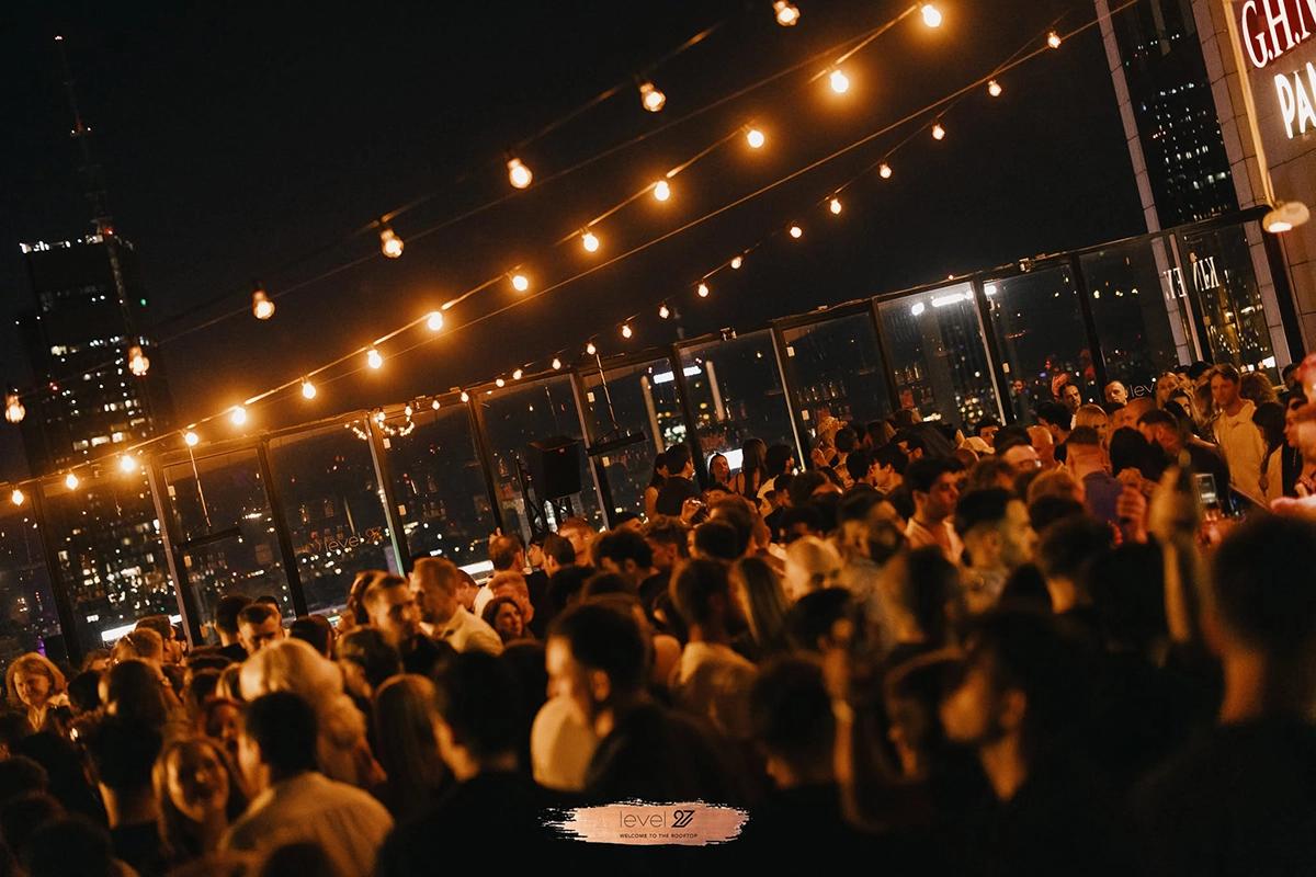 Vibrant night scene at level 27, a popular Warsaw rooftop nightclub with a crowd under string lights and a city skyline background.