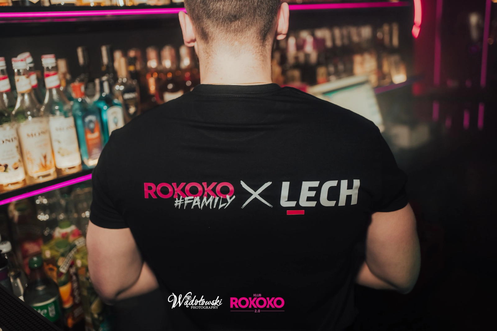 A close-up of the back of a staff member wearing a black t-shirt with the pink and silver "ROKOKO #FAMILY X LECH" logo, standing in front of a bar shelf filled with various spirits.