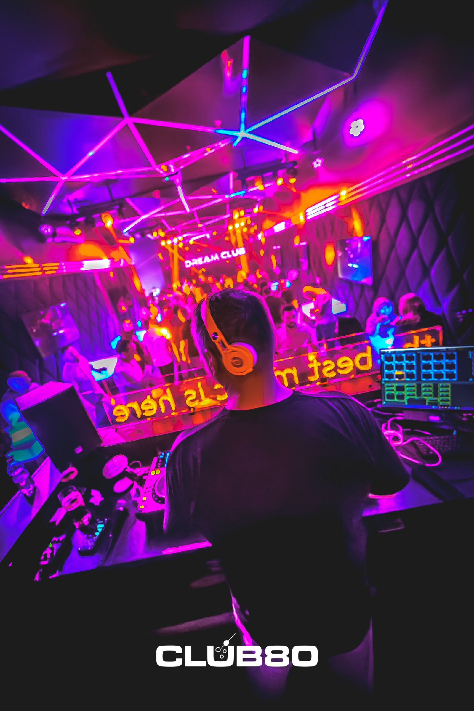 An over-the-shoulder view of a DJ wearing headphones, looking out at a packed dance floor illuminated by a spectacular geometric neon ceiling in shades of pink and blue.