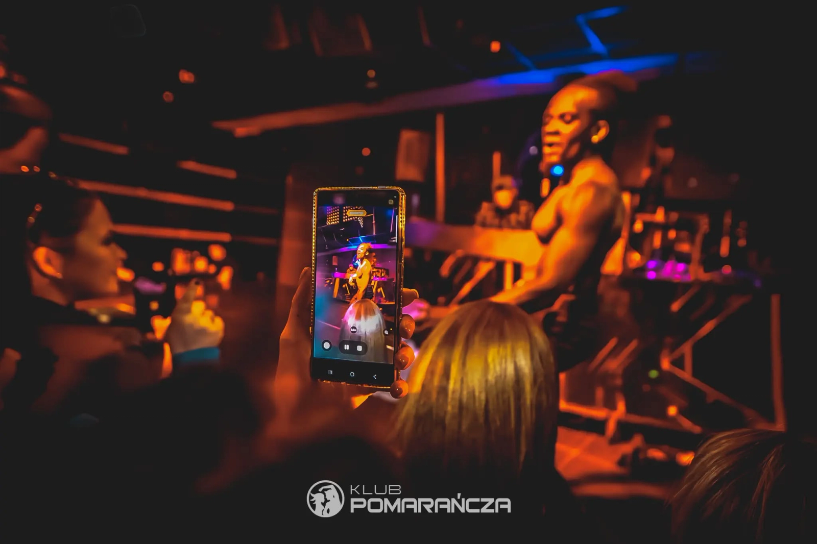 A clubgoer uses a smartphone to record a shirtless male dancer performing on stage under warm orange ambient lighting.