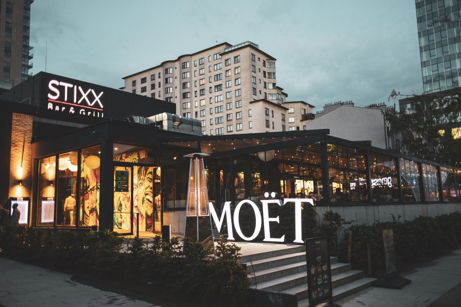 The exterior of STIXX Bar and Grill at twilight, featuring a glowing logo, a glass-enclosed patio, and a large illuminated MOËT sign on the front steps.