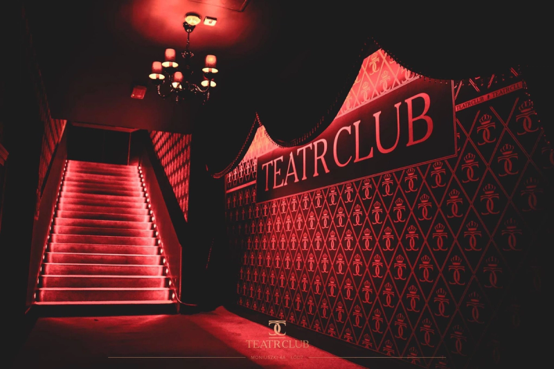 A dramatic, red-lit entrance staircase at Teatr Club in Łódź, featuring a branded wall with a repeating gold logo pattern and an elegant classic chandelier.