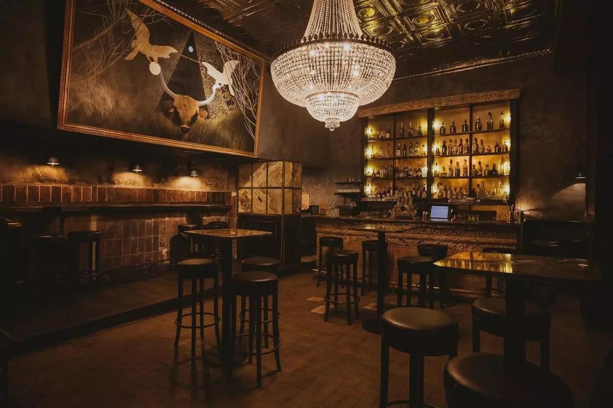 A dark, empty speakeasy lounge with a glowing back bar, a massive crystal chandelier, high-top tables, and a large surreal painting of a bull skull on the wall.